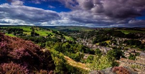 A view of the countryside in the sunshine with fields, trees and distant hills, with the town down in the valley. The sky is blue, but with some clouds threatening rain.