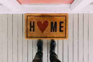 Looking down at a doormat in front of a closed door.  Home is written on the doormat with a heart instead of the letter O.  You can also see the bottom of a man's legs, wearing jeans and trainers, standing in front of the mat.