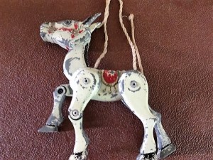 A well worn Muffin the Mule with four strings. He is white, with a black mane and ears and is wearing a red halter and saddle. 