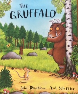 The cover of The Gruffalo written by by Julia Donaldson and illustrated by Axel Sheffler.  It shows the large smiling Gruffalo in the woods and a little mouse walking along a path.