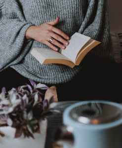 A woman's hand holding open a paperback book so that she can  read it.  In the foreground is a coffee pot and  plant on a table.  