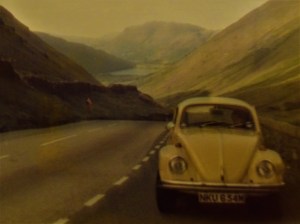 The yellow Beetle, parked on the side of the road with a view of mountains and a lake behind