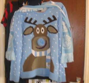 A Christmas jumper in the colours of the Manchester City football kit (light blue and white) on which is a brown reindeer with a blue pompom for a nose and wearing a Manchester City football scarf .