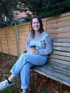 A photograph of Ellie sitting on a bench besides a garden fence with a takeaway cup of coffee.  She is smiling at the camera.  She is wearing blue dungarees over a grey jumper and trainers.  There are autumn leaves on the grass around her.