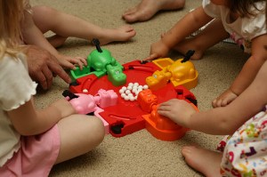 A photo of children crowded round a game of Hungry Hippos.  Four brightly coloured plastic hippos surround the hollow containing white balls.  The children have to catch the balls with the hippos mouths, opened by a lever on their tails.  