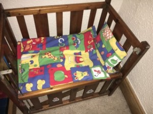 Linda's toy cot made from polished wooden bars, with the side dropped. Inside there is a red, yellow blue and green toy duvet and pillow with pictures of an apple, bear, car, ducks etc. used to teach the alphabet.
