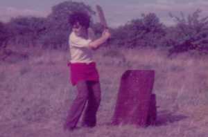 Curly haired Tony as a young teenager with his cricket bat raised ready to hit the ball, in front of a homemade wicket made from a piece of metal leaning against a post.  He is wearing a white shirt with sleeves rolled up, brown flared trousers and a red jumper tied round his waist. 