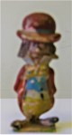 A brightly painted plaster figurine, with a large red bowler hat, long hair, moustache, red jacket, short blue waistcoat and yellow trousers held up by braces.  He wears long pointed shoes like a clown's, with feet pointed outwards and his hands are behind his back.  