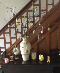 Linda's Easter display. A tall vase on a table contains the bare branch decorated with pink, blue and gold eggs. Around the vase are a selection of china hens of differing sizes and a china teddy bear.