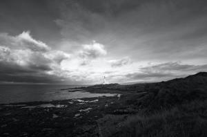 A dramatic black and white photograph of a rocky shoreline with a lighthouse in the distant horizon, lit by a sunset through the clouds.