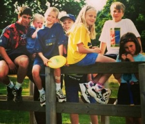 Seven children from a toddler to a teenager, wearing bright summer clothes, sit on a fence in front of grass and trees.  One of the boys is holding a frisbee. 