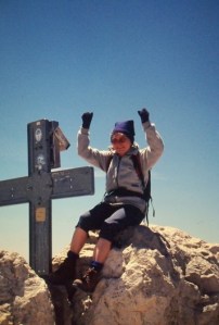 Margaret sitting in the sunshine next to a cross marking the top of the mountain against a deep blue sky.  She has her hands raised in victory. 