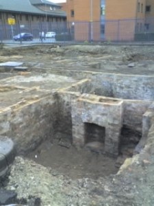 The excavations showing a fireplace in a cellar in a brick-built square room, part of a street of similar houses. Modern university buildings are visible in the background, behind a high fence.