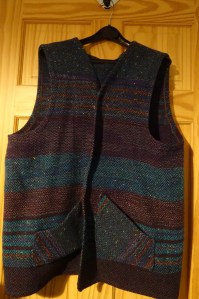 Margaret's long waistcoat is hanging on a coat hanger on the back of a wooden door.  It has stripes of diffferent widths and colours, including purples, browns, turquoise and green, and has vertical patch pockets.