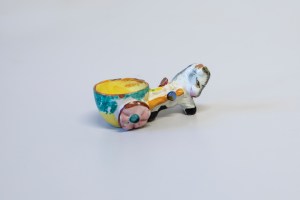 The donkey egg cup is brightly painted.  The cart is round, bright yellow inside with green leaves on the outside.  The wheel is brown with pink spokes.  The donkey is white with black hooves and looks like it is smiling.