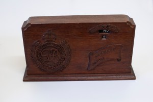 A wooden date display calendar with a carved royal crest containing the letters GR above which is a crown.  There are carved wings above the display of the month and date, which can be changed by a dial on the back.