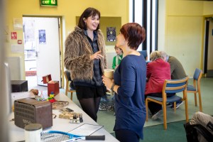 Jolene and a visitor are standing by a table displaying a range of items. In the background people are sitting round a table busily chatting.