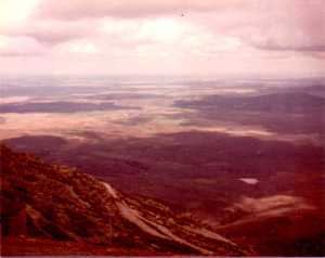 In the foreground, the side of the mountain has a rocky path with sparse grass.  The view beyond shows how high they'd climbed, with fields and trees stretching to the horizon, in patches of sunlight. 