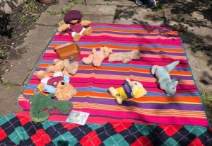 Five teddy bears and two green dragons are lying on their backs on a rainbow striped picnic rug, on top of flagstones. 