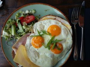 An inviting looking breakfast of layers of toast, ham, thinly sliced cheese topped with three fried eggs sprinkled with pea shoots.  There is a side salad of chopped lettuce, cucumber and tomato.  Besides the plate, a shiny silver knife and fork are ready to be picked up.