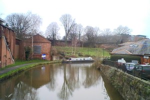 The photograph shows reflections of a red brick old building along the left hand side of the canal, and of the bare branched trees behind and further along round a bend.  There is a moored narrowboat painted white and green just before the bend.  On the right hand side is an old stone wall. 