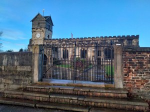 A view of the unusual church through closed wrought iron gates.  The clock tower has a wooden, roofed building on the top with a golden weather vane.  The steps in front of the railings and the paths leading between lawned areas leading to the church are shiny and wet following the rain.  But the sky is a lovely shade of bright blue.