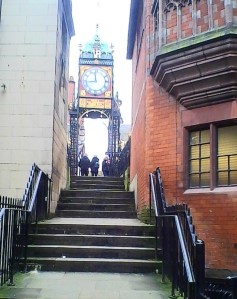 The gold surrounds of the ornate clock shine in the winter sunshine at the top of three flights of four steps, on the narrow walk way between two buildings. One of the buildings is red brick with a distinctive window overhanging the steps.  The other is white stone.  There are wrought iron railings leading up to the top of the first flight of steps.
