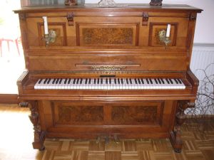 An old-fashioned upright piano with inlaid walnut and ornately carved legs.  There are two brass candlesticks with partially used white candles on either side. 