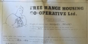 A photograph of Free Range Housing Cooperative share certificate from 1984, with a cartoon drawing of a hen sheltering a house. 