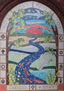 The mosaic is in the arch of a former doorway in the red-brick building. It shows sky with moon and stars under which there is a setting sun.  From the horizon, runs a blue river full of orange fish across fields and paths.  Two trees mirror the arch on either side of the mosaic, with a dove holding an olive branch Flying besides it.  There are daffodils and lilies of the valley in the foreground.  At the top of the arch the words HOPE, LOVE, PEACE, JOY are written in mosaic, and around the border there are symbols of different religions.  At the bottom, it says CREATED 2016
