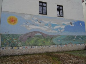 The mosaic is along the whole side of a building, underneath two windows.  It shows a blue sky with a bright sun and clouds, with geese and doves flying from left to right.  There is a stone arched bridge on land covered with brightly coloured flowers, stretching as far as the eye can see alongside a lake with water lilies and dragonflies.