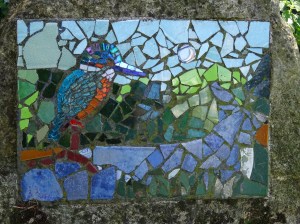 In the mosaic, the kingfisher is standing on the branch of a tree by the river. There are trees and hills in different shades of green behind it, against a pale blue sky. 