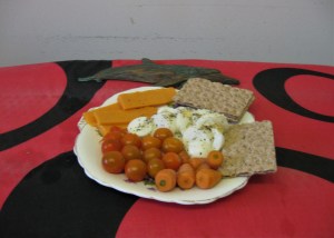 A scallop-edged white plate containing an arrangement of cherry tomatoes, sliced hard-boiled eggs, slices of Cheddar cheese and four crispbreads. the plate is on a bright red tablecloth with swirls of black, with a copper ornament of a fish behind it.  