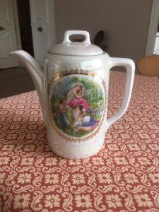 The coffee pot is white with an oval picture of a girl in a classical pose, sitting by water in a forest.  The picture has a fancy gold border. 