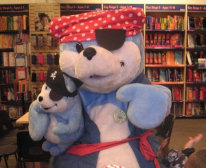 The large, blue and white bear is wearing a red kerchief with white spots, and a patch over one eye.  The bear is holding a smaller cuddly toy version wearing a pirate's hat.  You can see books on the library shelves in the background.