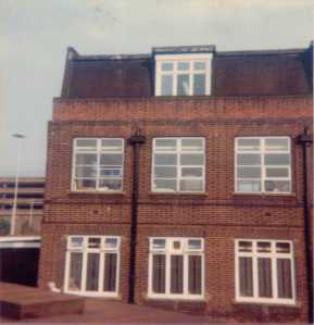 A three storey red brick building with six large windows on the ground and first floors, and a further full-sized window set back in the roof.  In the background, you can see a car park which is probably behind some railway tracks.