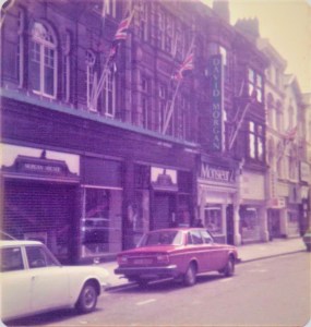 The shop frontage of the Department store has flag posts with Union Jack flags at an angle.  There are 1970s cars parked outside.