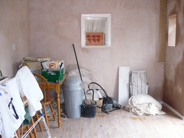 The kitchen walls have been re-plastered and most of the furniture removed. There are fittings, a hoover, a mop and bucket and a table with boxes of tiles on it.