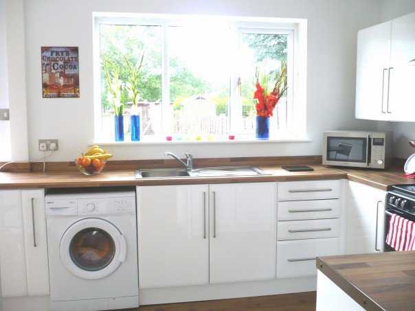 The finished, gleaming white new kitchen with wooden surfaces and all "mod cons". The sun is shining through the window and there are gladioli in a vase on the windowl edge. 