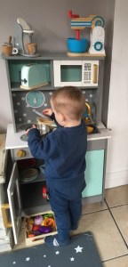 One great nephew standing in front of  the toy kitchen, putting a lid on a pan.  On shelves behind the cooker there is a  toy toaster, microwave, food mixer and coffee maker.