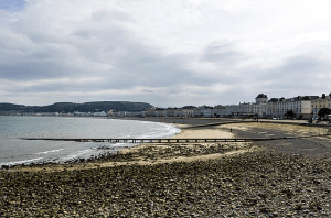 A sweeping view of Llandudno beach, with pebbles in the foreground and the sun shining on a gentle sea with waves lapping the sandy shore in the middle of the photograph.  The bay is lined with the traditional white-painted seaside hotels and there is a view of the Little Orme at its end.