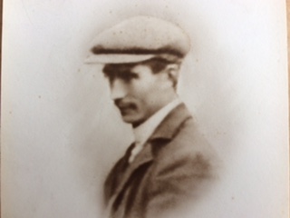 A photograph showing a head and shoulders side profile  of Shane's grandfather as a young man.  He is smartly dressed, wearing a jacket, collar and tie and a large peaked cap.  His hair is short, he has bushy eyebrows and neatly trimmed moustache.