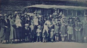 A photograph of a group of around thirty adults and children wearing coats, hats and caps.  They are standing in front of an old coach (a charabanc) with a cloth roof.