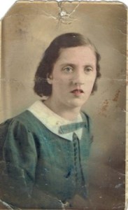 The young white woman, with shoulder length brown hair,  is looking slightly past the camera to the left.  The dress has a white collar and has been coloured bluey green. 