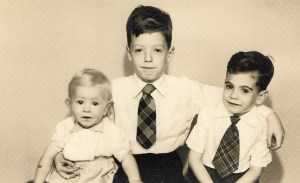 The eldest brother is in the centre with his arms round his two younger brothers.  The older boys have dark hair and are wearing white shirts and checked ties, the youngest boy is a toddler.  He  has fair hair and is wearing a romper suit.   