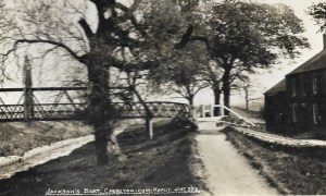 The caption on the postcard reads Jackson's Boat, Chorlton-cum-Hardy.  There is an iron bridge crossing the river to the left, and the public house called "Jackson's Boat" is to the right of the path running alongside the river.
