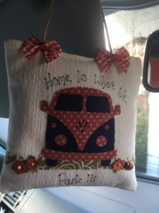The text on the lavender bag says "Home is where we park it". There are red and white bows on the string from which it is hanging, an applique of a classic VW campervan in blue and red polka dots and appliqued flowers on the grass beneath it.