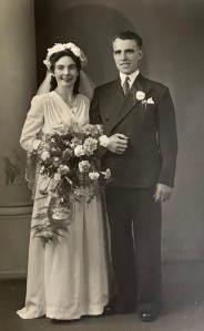 Shane's parents are smiling at the camera in their wedding photograph. His mother is wearing a headdress, veil and long traditional white wedding dress. She is carrying a very large bouquet of flowers from which hangs an imitation horseshoe. Shane's father is standing up straight, wearing a shirt with a high collar, tie, suit with a double breasted jacket, a neatly folded handkerchief in the breast pocket and a carnation in the buttonhole. They are linking arms, his father's right arm is folded across his waist, and his left arm is hanging by his side. Both hands are visibly large.