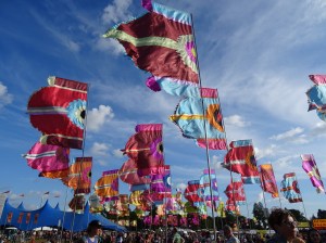 A view of the festival site with large brightly coloured flags on poles  waving in the breeze with a bright blue sky in the background.  A marquee is visible in the distance and crowds of people.