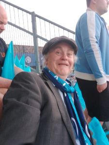 A photograph of Tony wearing a traditional flat cap and sports jacket and his Manchester City Supporters "match scarf"
